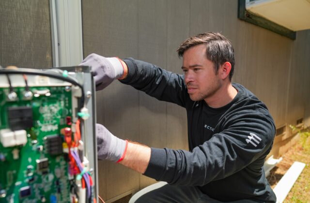 A technician wearing a long-sleeved black shirt and protective work gloves carefully works on an open electrical control board of an outdoor heat pump against a house.