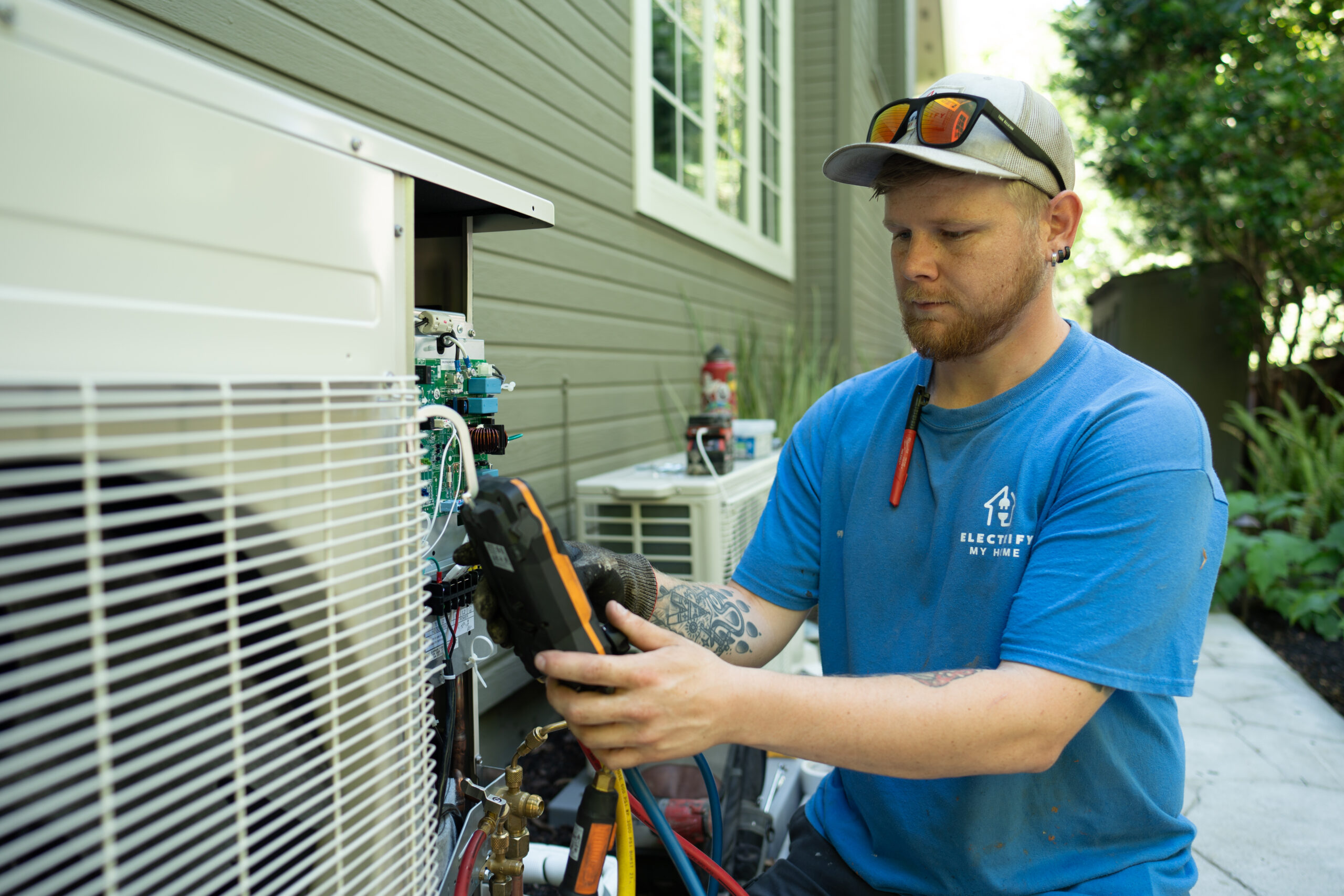 A technician wearing a blue t-shirt and a grey baseball cap works on the outdoor unit of a residential heat pump system.