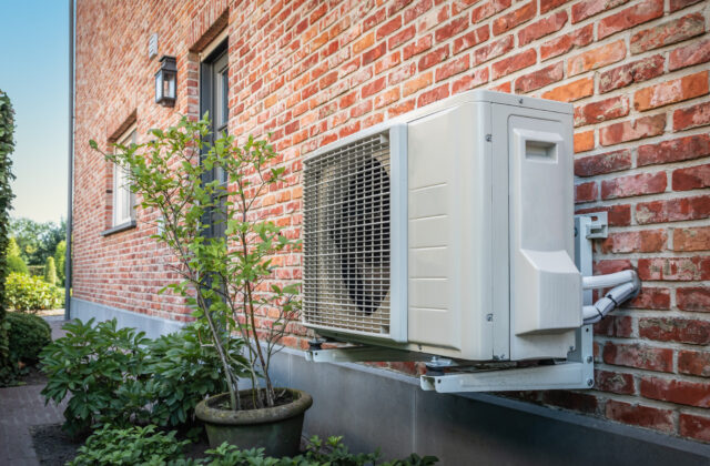 A white heat pump outdoor unit mounted on wall brackets against a red brick house, with a potted shrub and garden plants beside it.